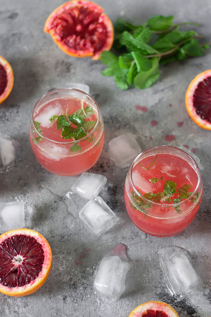 two blood orange cocktails on dark grey board, surrounded by blood orange segments, mint and ice cubes