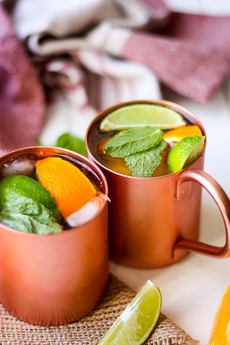 two copper mugs of citrus ginger dark and stormy cocktails on white board, with red and white tea towel in background