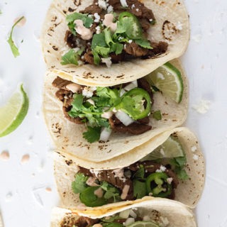 four steak tacos lined up topped with jalapeno, cilantro, onion and cojito