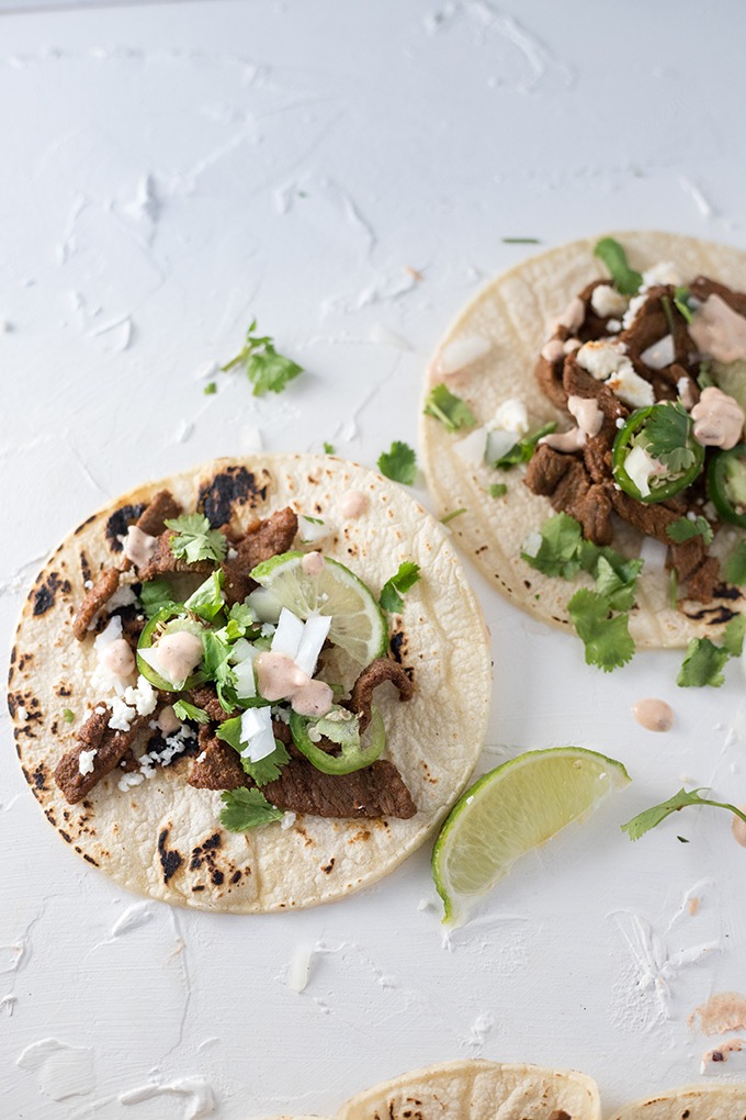 two steak tacos on a white board, topped with queso, cilantro, lime wedges and white onion