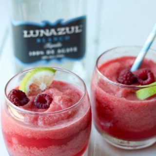 two tumblers of raspberry margaritas, second with blue straw in front of tequila bottle