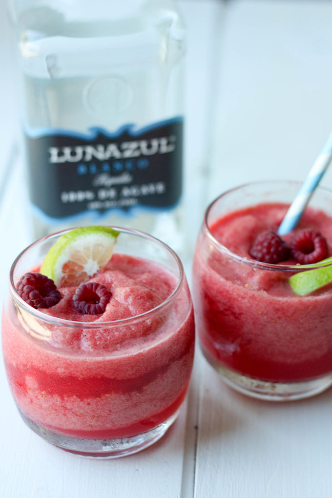 two tumblers of raspberry margaritas, second with blue straw in front of tequila bottle