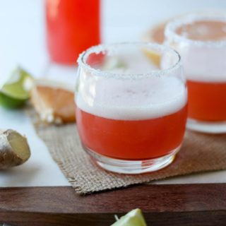 one grapefruit margarita on marble/wooden board, lime and orange wedges in front, grapefruit juice in background