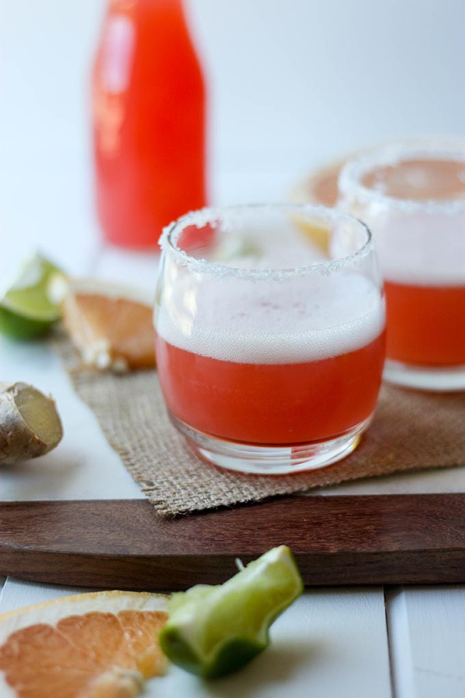 one grapfruit margarita cocktail on a marble board, lime and grapefruit wedges in foreground, bottle of grapfruit juice in background