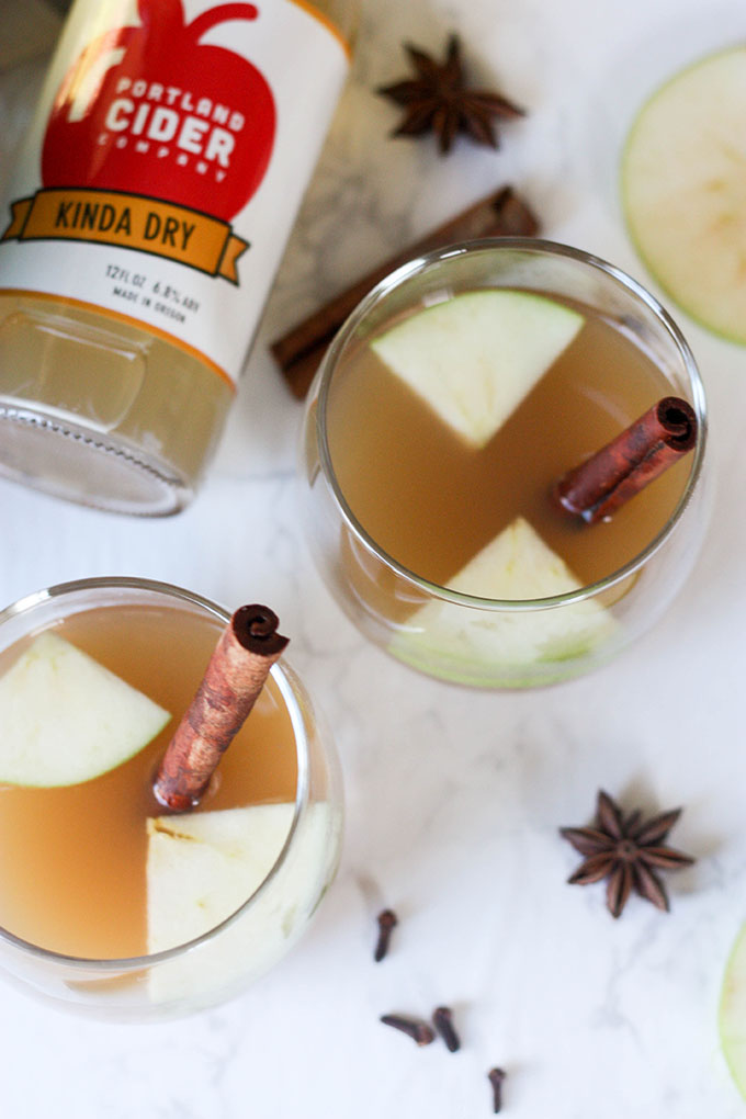 two glasses of hot spiced apple cider with cinnamon quills, bottle of cider behind glasses