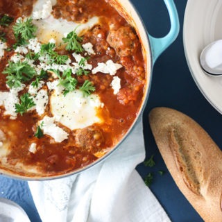 blue dutch oven with meatball spicy shakshuka, on white cloth and blue board, loaf of bread next to dish