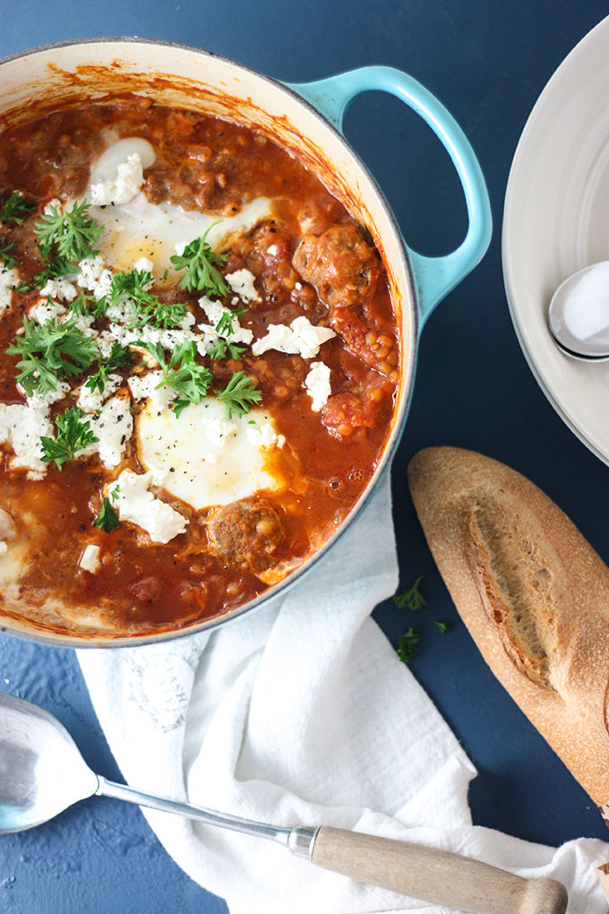 blue dutch oven with meatball spicy shakshuka, on white cloth and blue board, loaf of bread next to dish