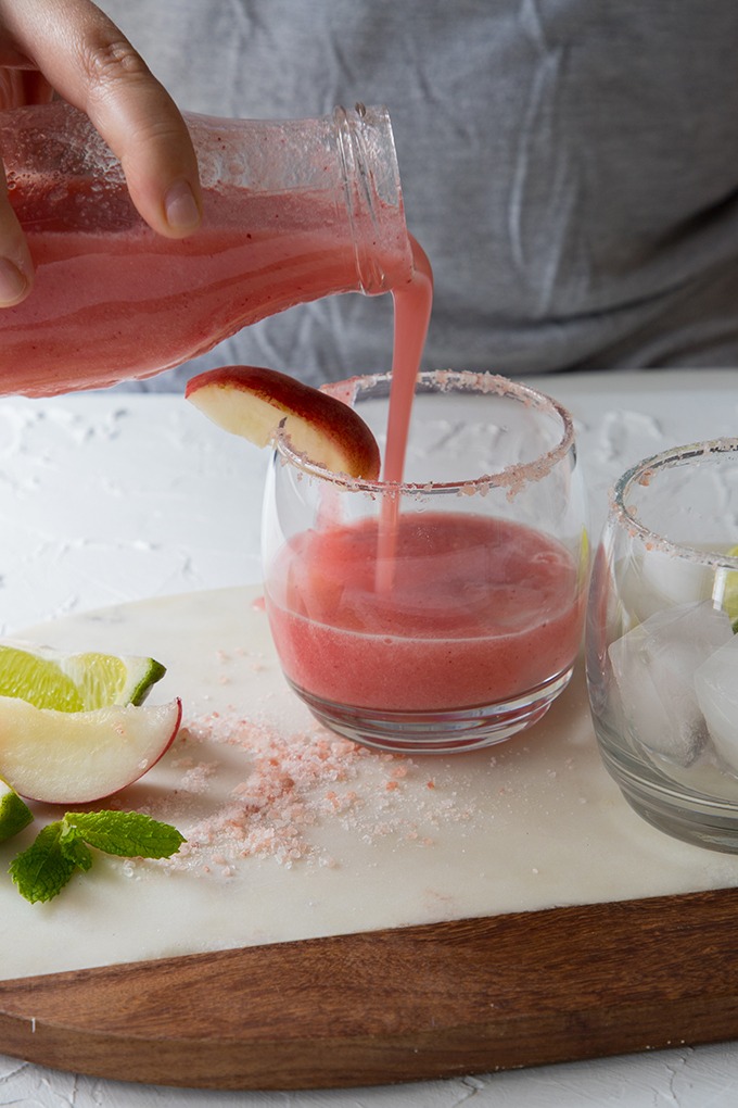 person standing behind a table, pouring the peach margarita into two clear glasses that are on a white marble board