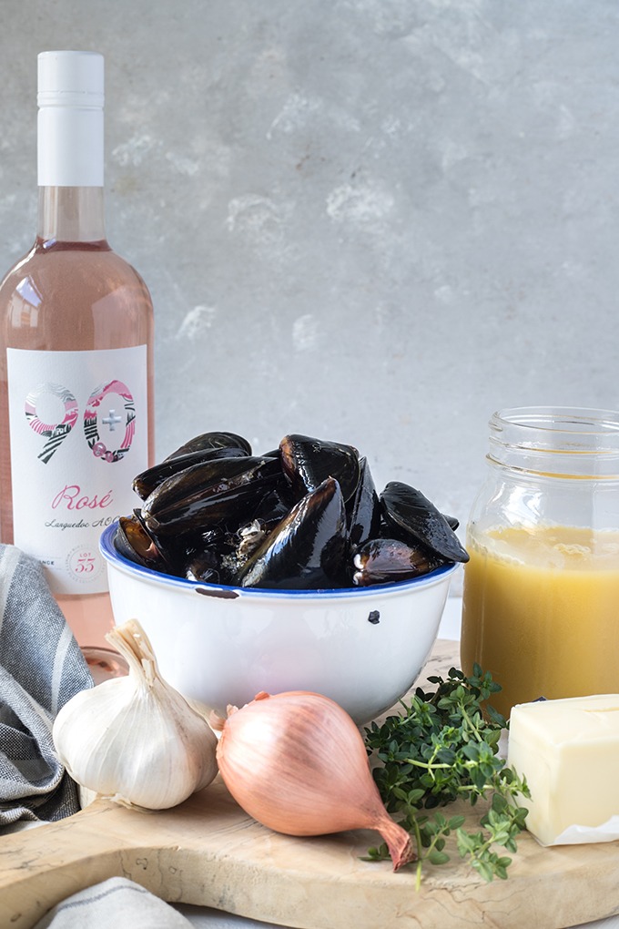 rosé wine mussels recipe ingredients on wooden board, left to right. rosé wine, mussels, chicken broth in jar, stick of butter, fresh thyme, one shallot and garlic clove