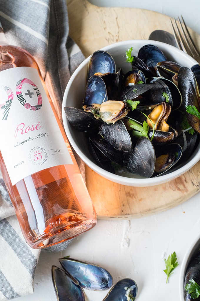 white bowl of wine mussels on wooden board right of a bottle of 90+ cellars rosé wine