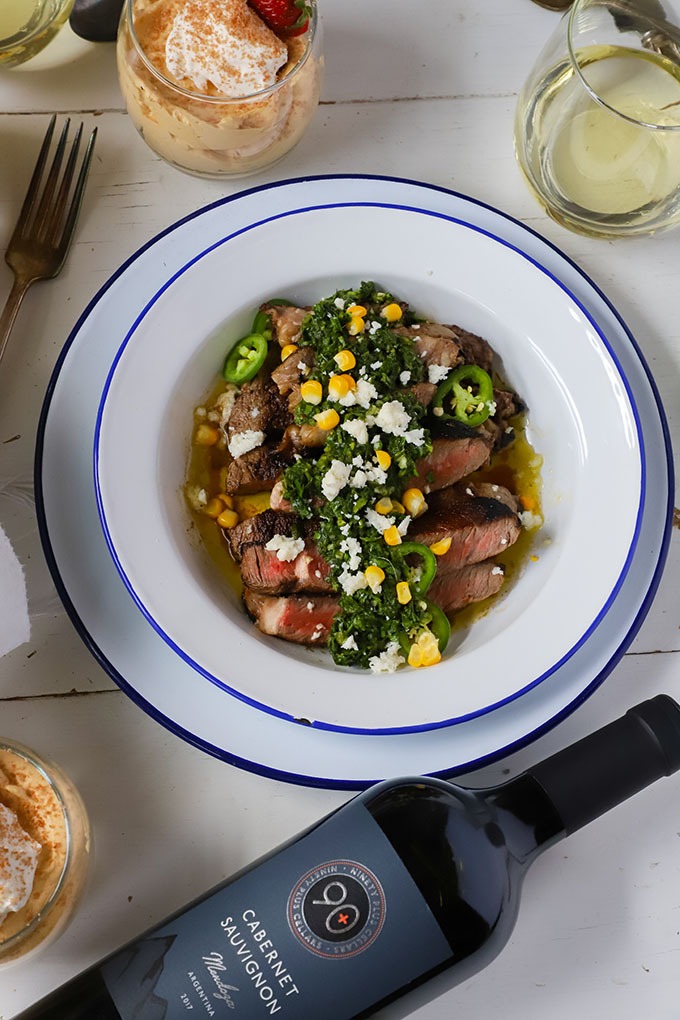 steak with chimichurri on white and blue enamel plate placed next to bottle of red wine