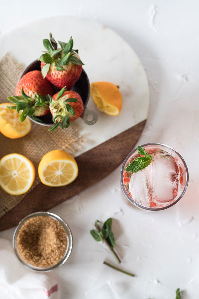 overhead of strawberry mojito recipe, marble wooden board topped with lemons and strabwerries, mojito in tall glass