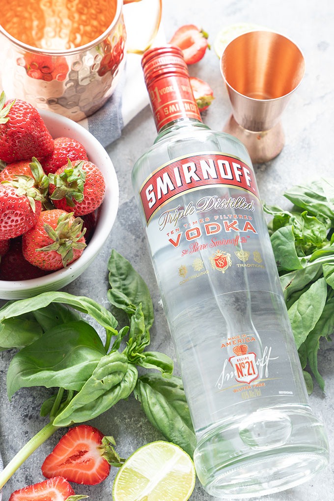 Smirnoff vodka bottle laying flat surrounded by strawberries, basil and copper measuring cup