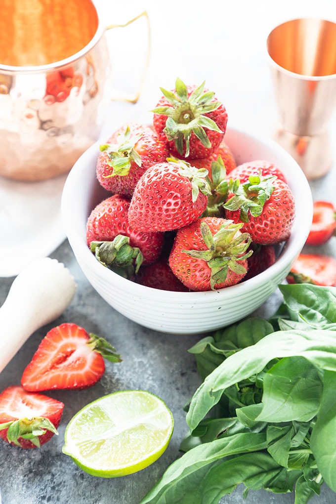 bowl of strawberries, lime wedges, basil, copper measuring jig and copper mug on grey board