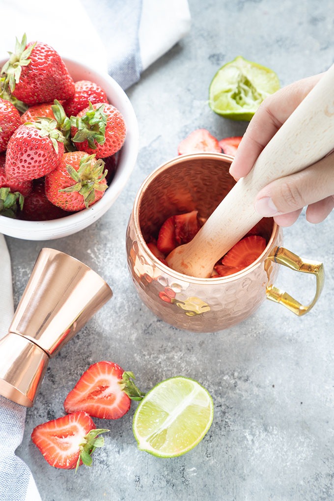 hand holding wooden muddler, muddling strawberries in a copper mug