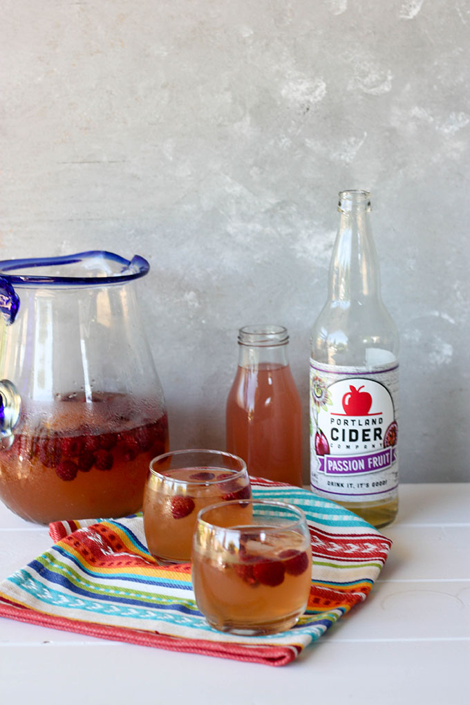 two small glasses of passion fruit cider punch on a colourful napkin. Pitcher, cider bottle and small jug of juice behind glasses