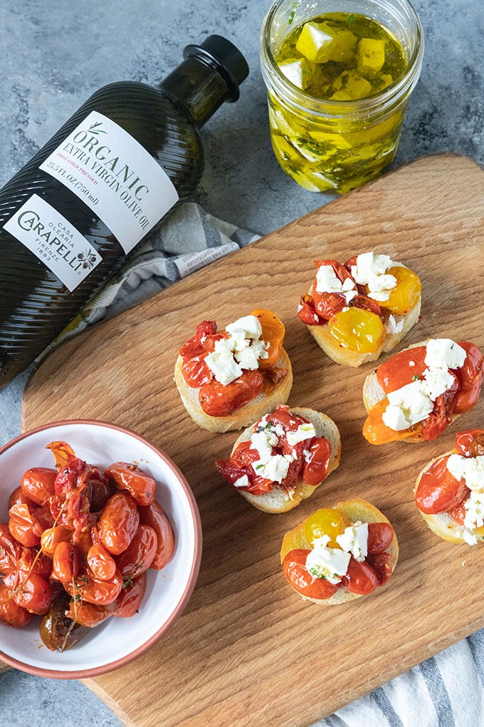 tomato crostini on wooden board with bowl of roasted tomatoes, olive oil and feta jar