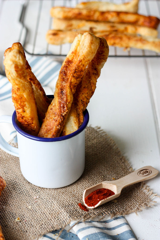 blue and white enamel mug on a hessian square. mug full of parmesan puff pastry cheese straws, extra straws on a wire rack in background